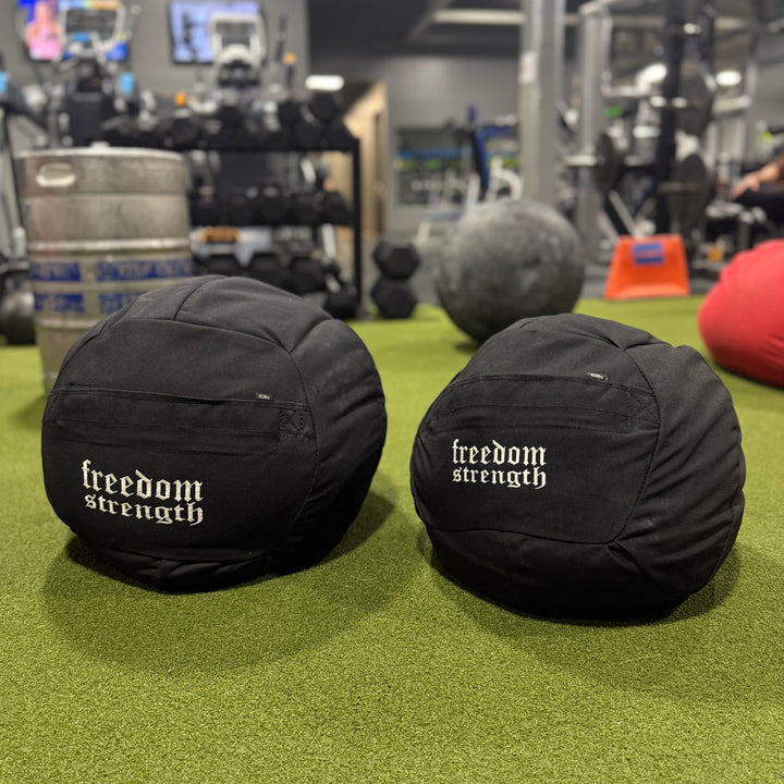 Two black atlas sandbag medicine balls with 'freedom strength' branding on a green floor in a gym setting.