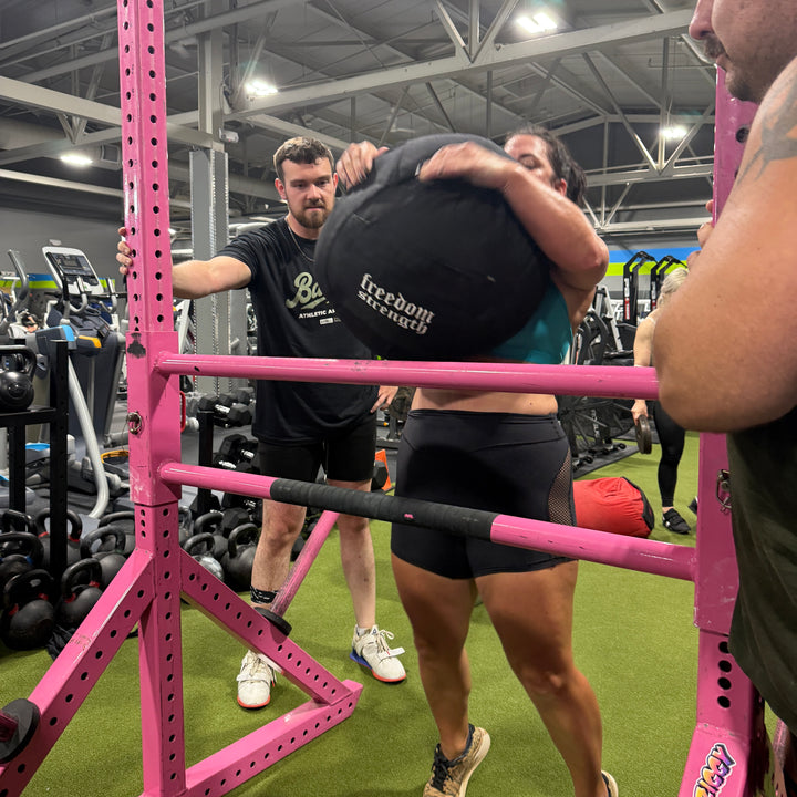 Person lifting a atlas sandbag medicine weighted ball with assistance in a gym setting