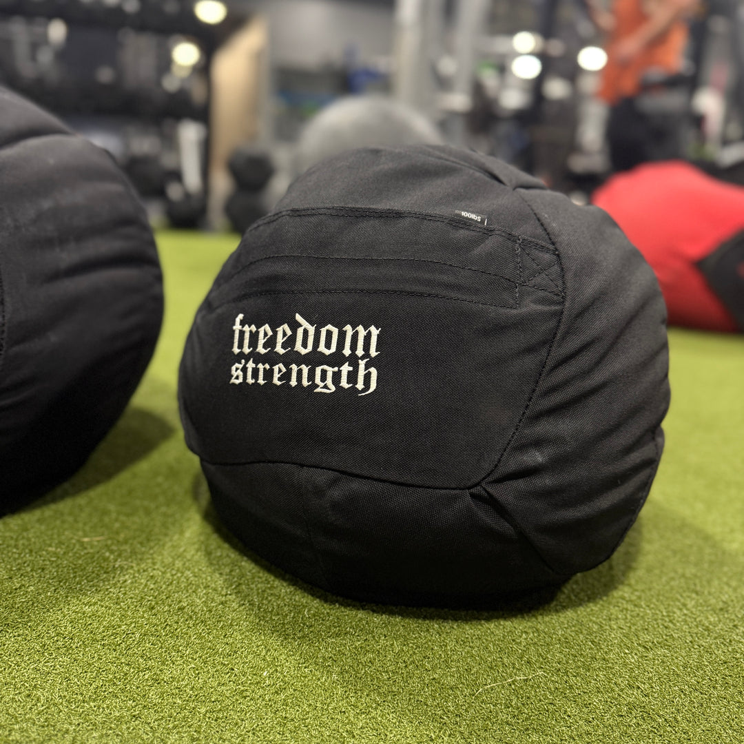 Black atlas stone sandbag medicine ball with 'freedom strength' text on a green floor in a gym setting